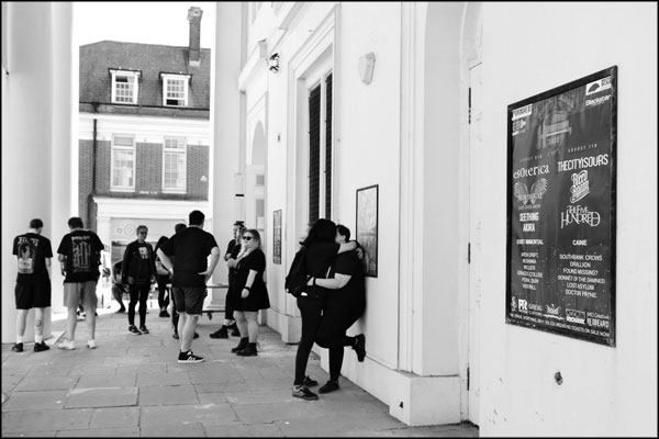 Outside the venue, attendees wait for the music, the event poster on the wall. Hammerdown Festival 2022, by Greig Clifford